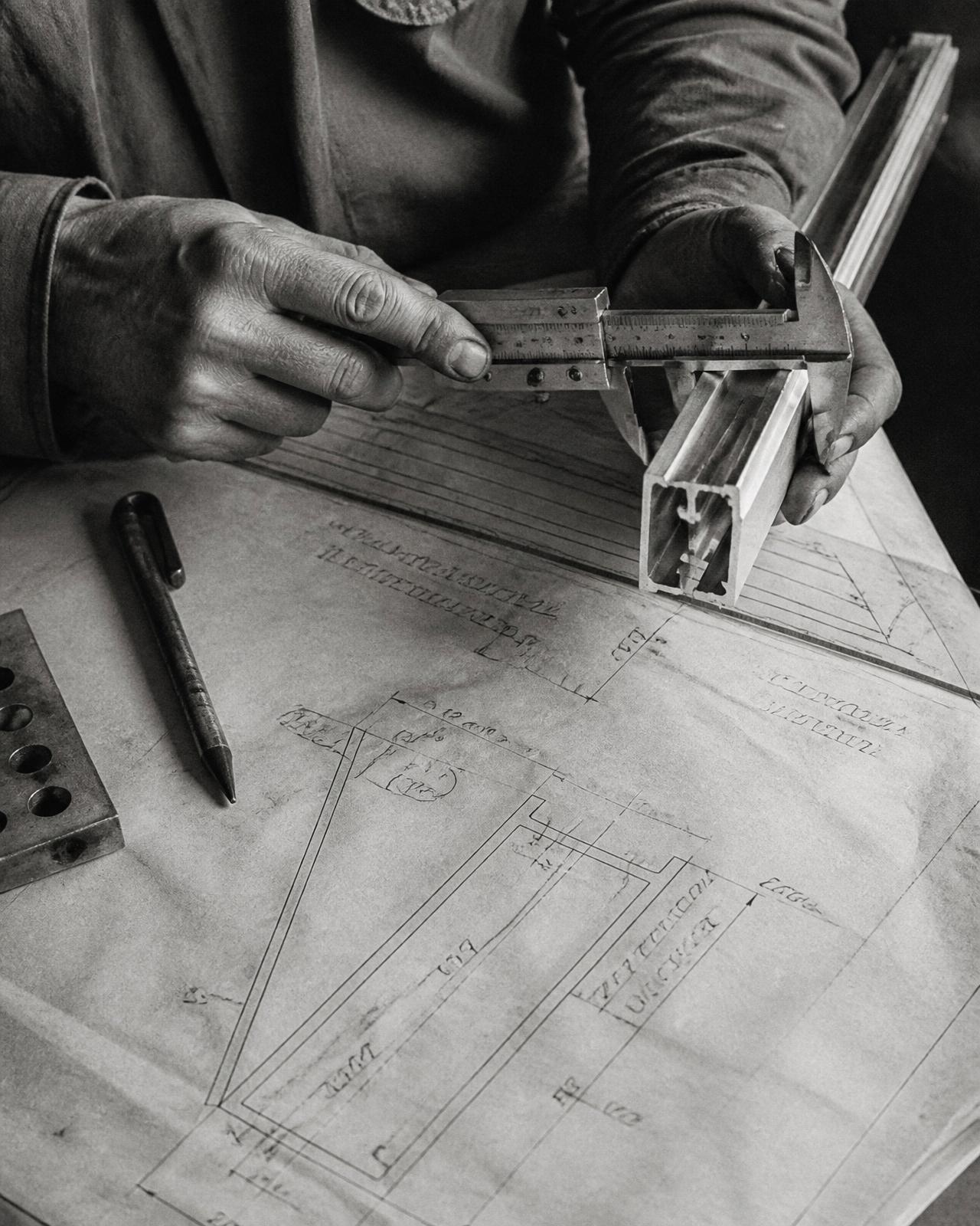 Mani al banco di lavoro con calibro su profilo in alluminio e disegni tecnici d'epoca, archivio anni Settanta